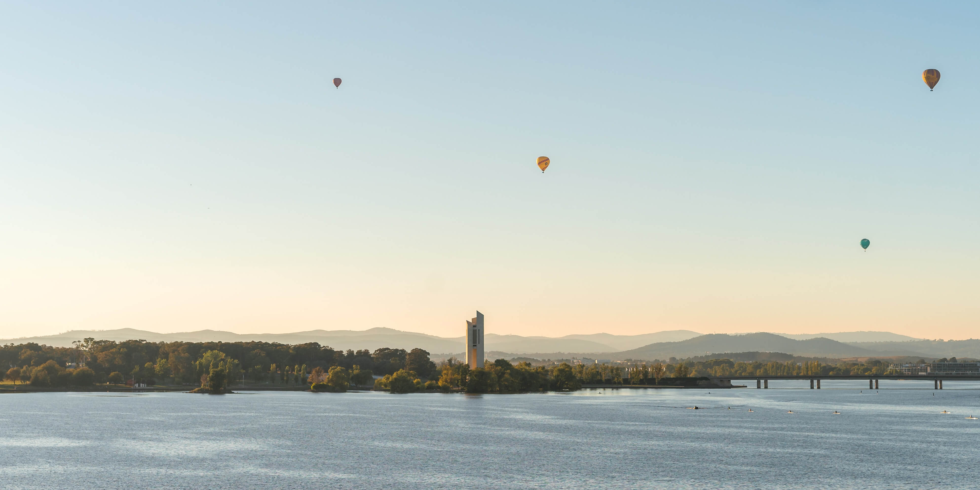 A lake in Canberra with a building in the middle and several hot air balloons at sunrise