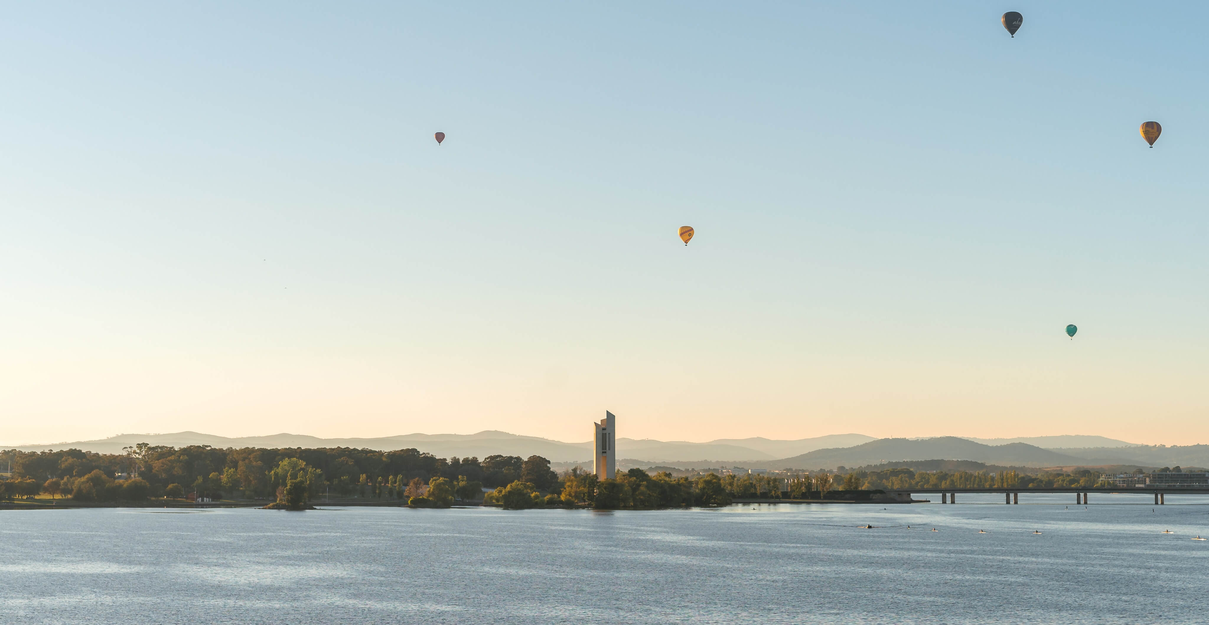 A lake in Canberra with a building in the middle and several hot air balloons at sunrise