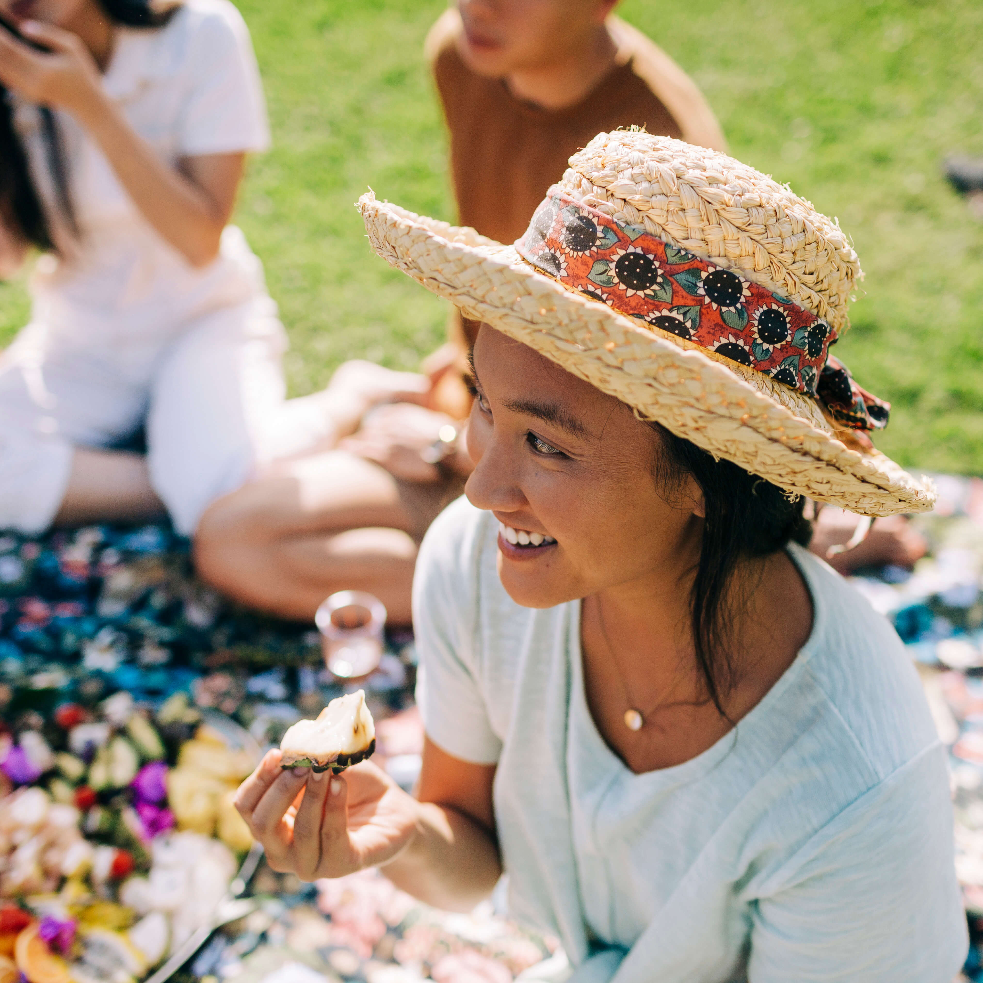 smiling woman with a straw hat on sitting at a picnic with food in her hand.
