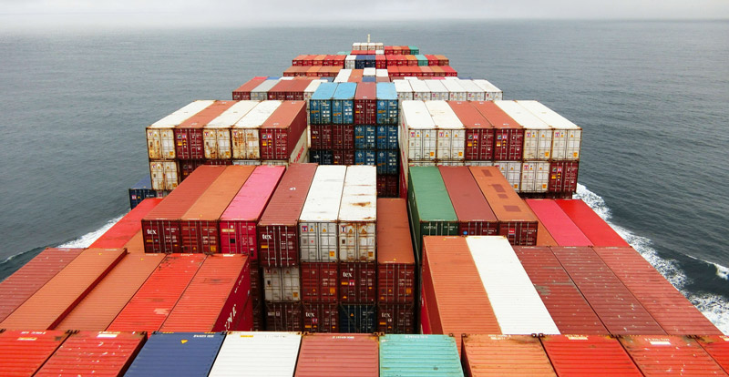 View from the bridge of a large shipping boat showing lots of stacked shipping containers. Somewhere in the middle of the ocean