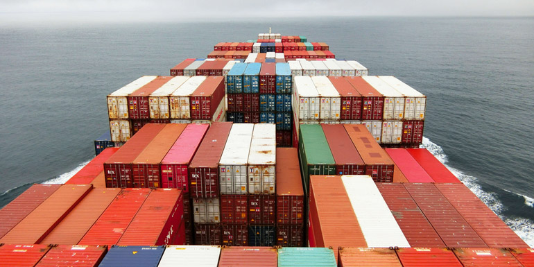 View from the bridge of a large shipping boat showing lots of stacked shipping containers. Somewhere in the middle of the ocean