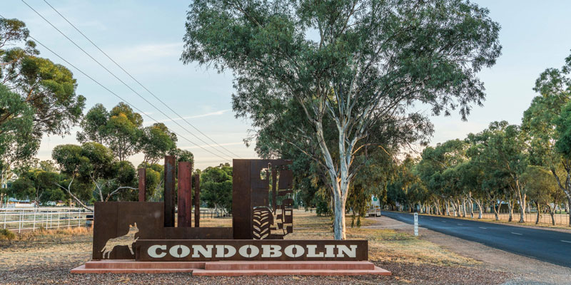 Image of a town sign that reads Condobolin that is made out of rusted iron, next to a road with a flood sign