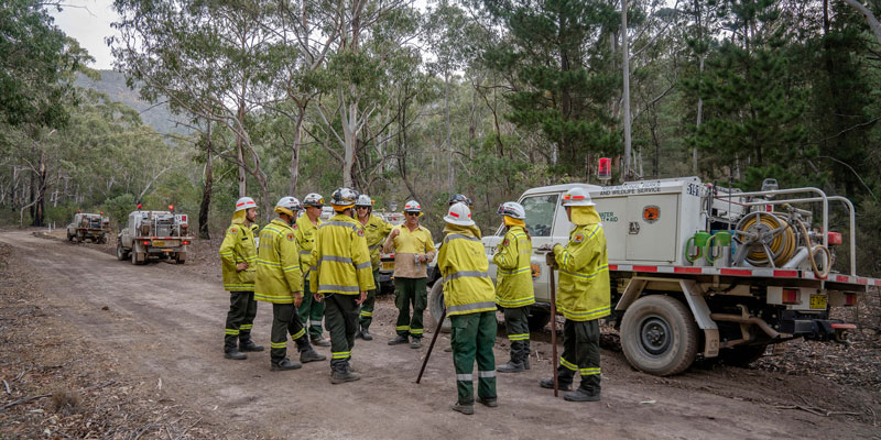 group of 12 firefighters talking to each other on a dirt road in the bush with a fires truck next to them