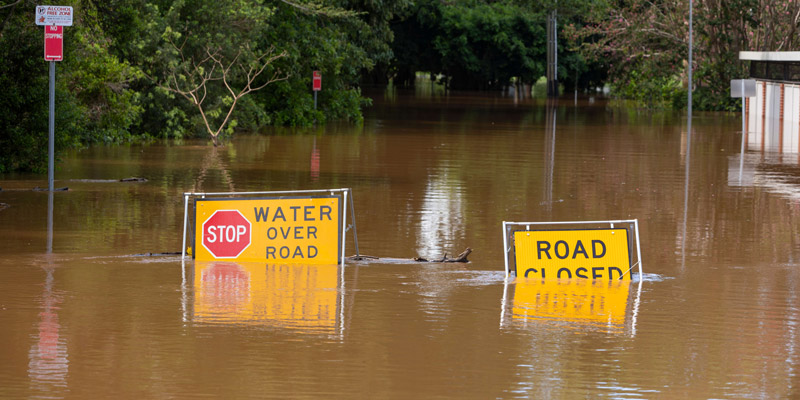 Close up of two signs saying 'water over road' and 'road closed' partially covered by flood water on a road 