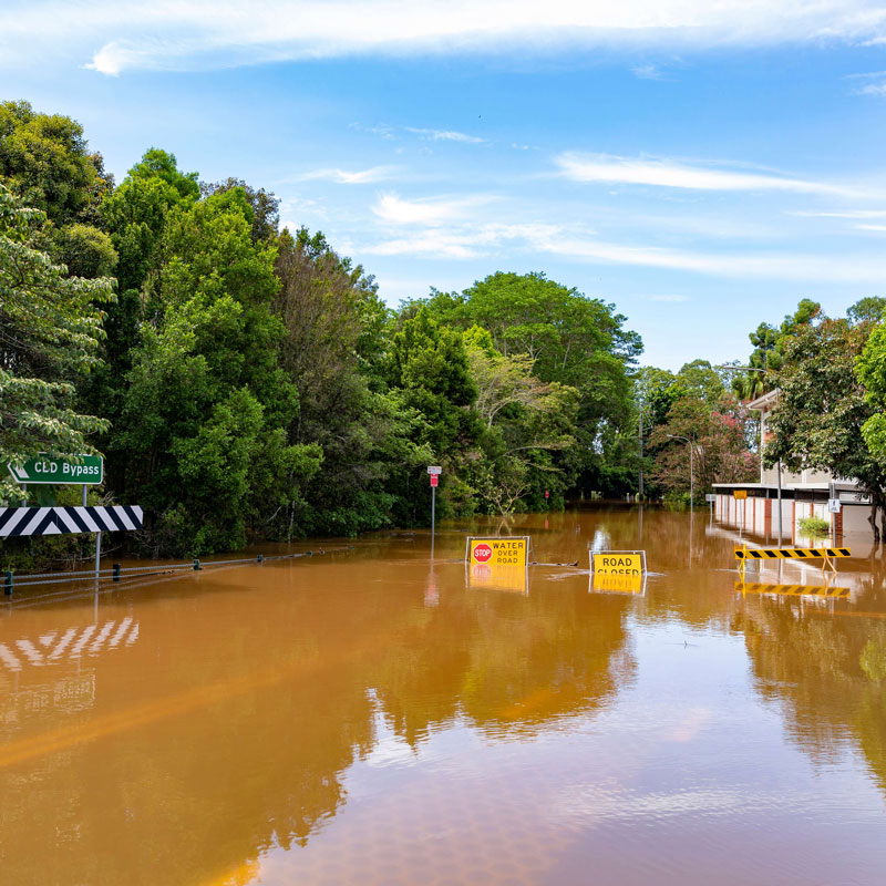 image of a flooded street with brown flood water, green trees and blue sky