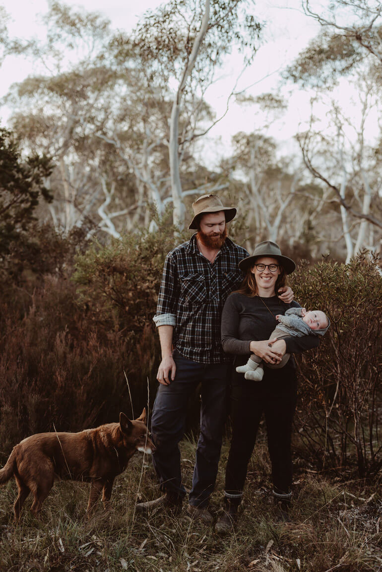 A couple holding a baby with a dog by their side stand in the bush 