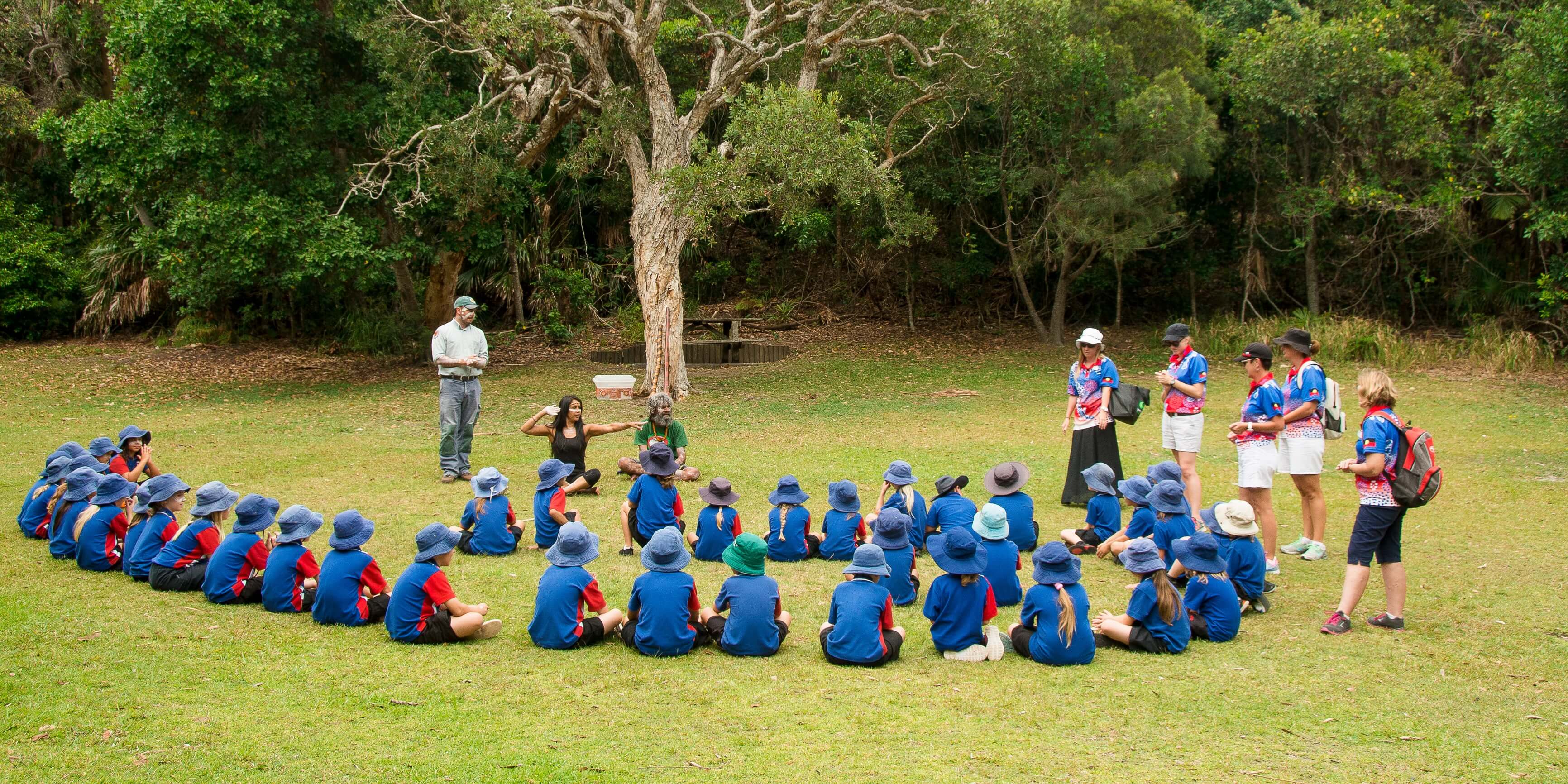 Group of school students seating on the grass listening to Aboriginal women. Image credit: David Young/DPIE