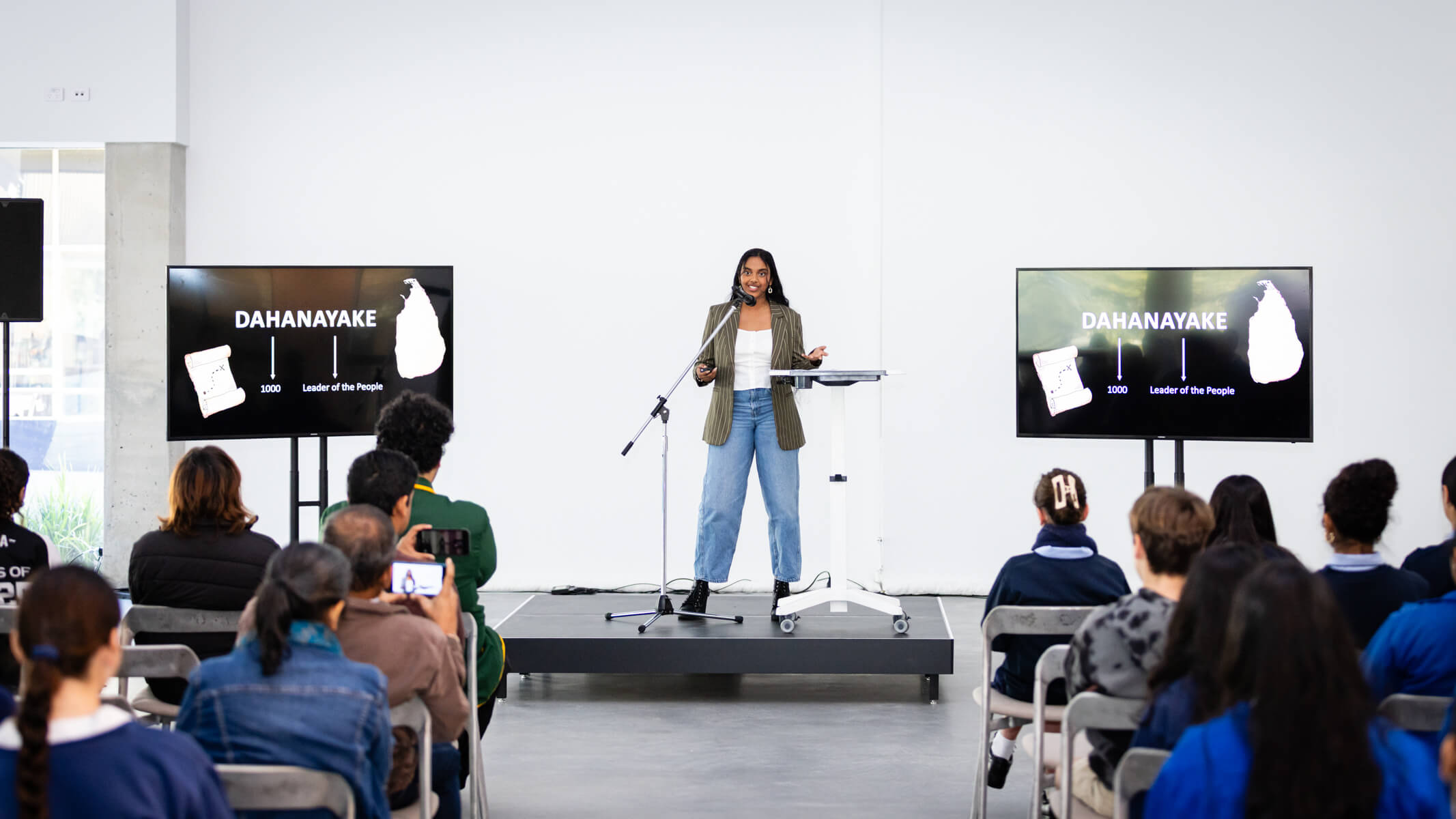 Yehansa Dahanayake stands on a stage in front of a room of high school students speaking into a microphone.