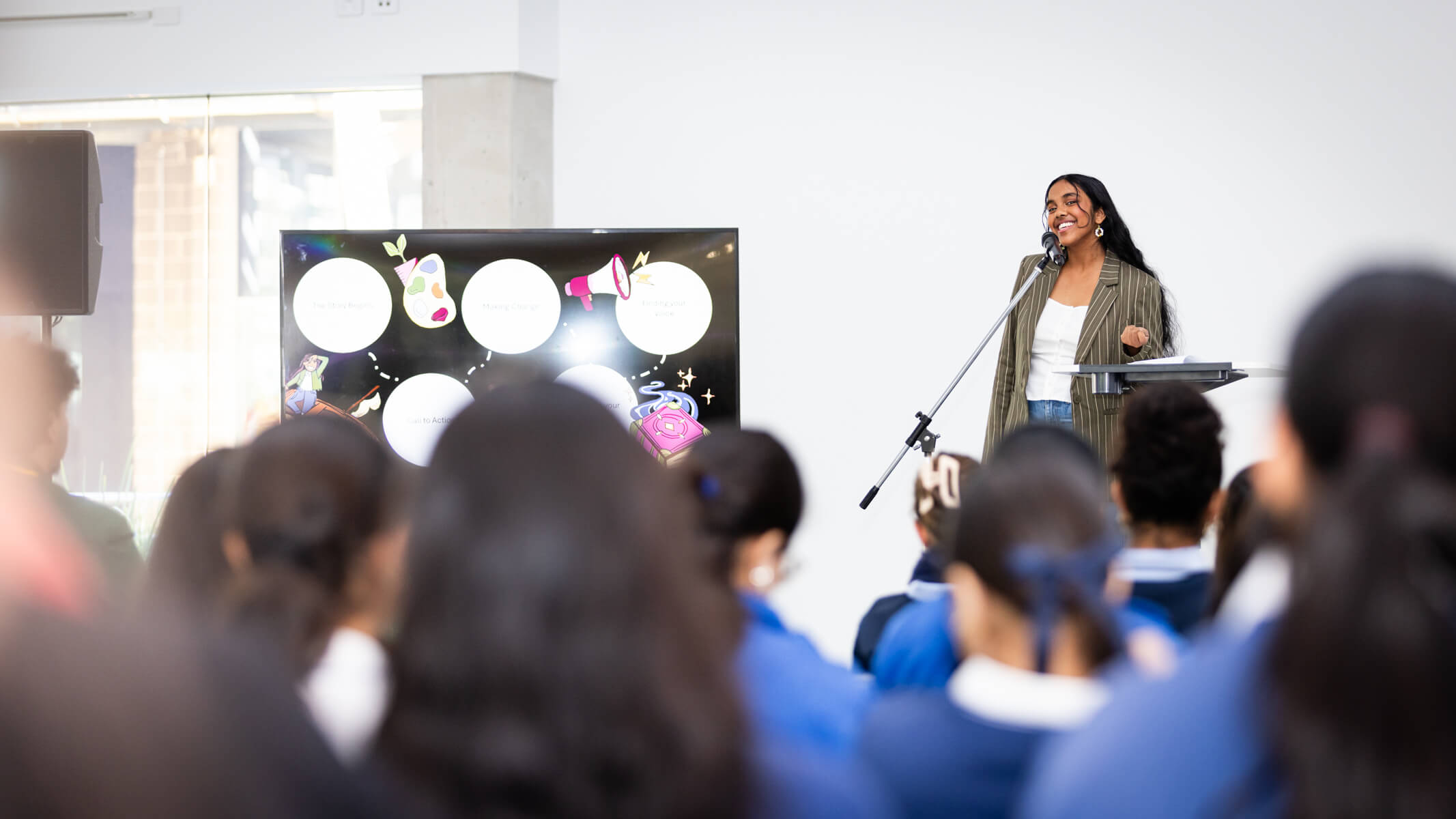 Yehansa Dahanayake, a youth climate justice advocate, artist, and researcher stands on a stage at the microphone speaking to a crowd of students.