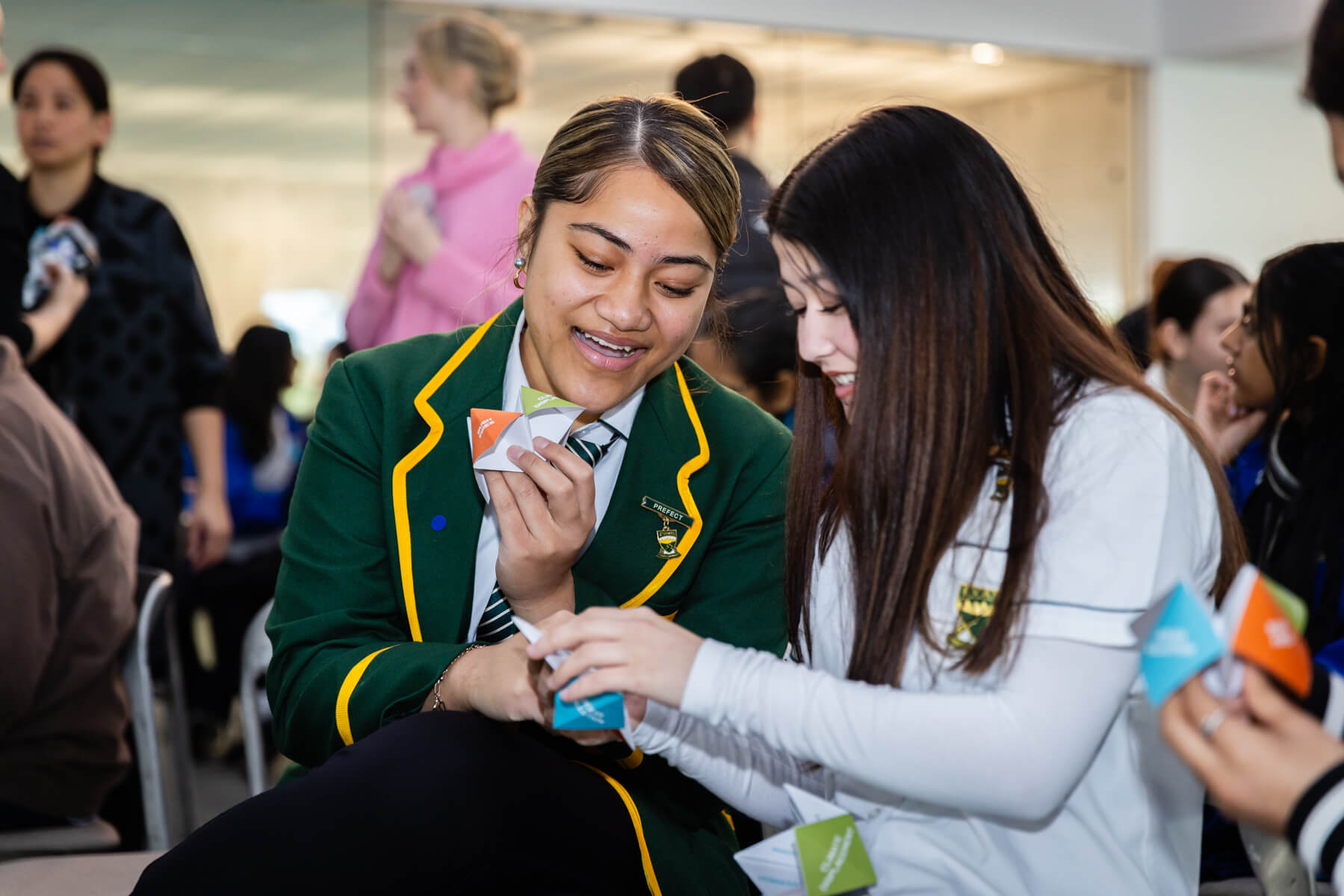 Two high school students sit amongst an audience smiling as they use paper chatterboxes.