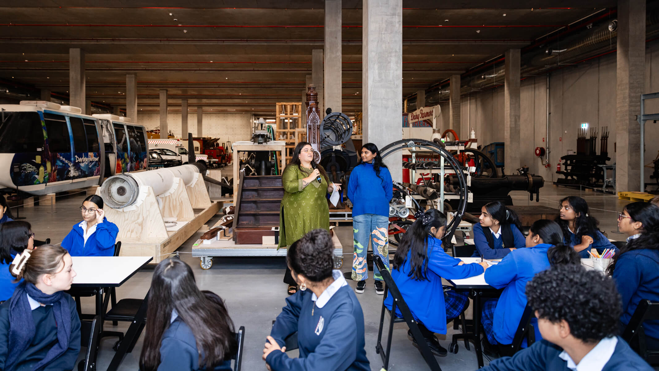 Two women speak to students sitting at tables within the Powerhouse Castle Hill Very Large and Complex Object storeroom.