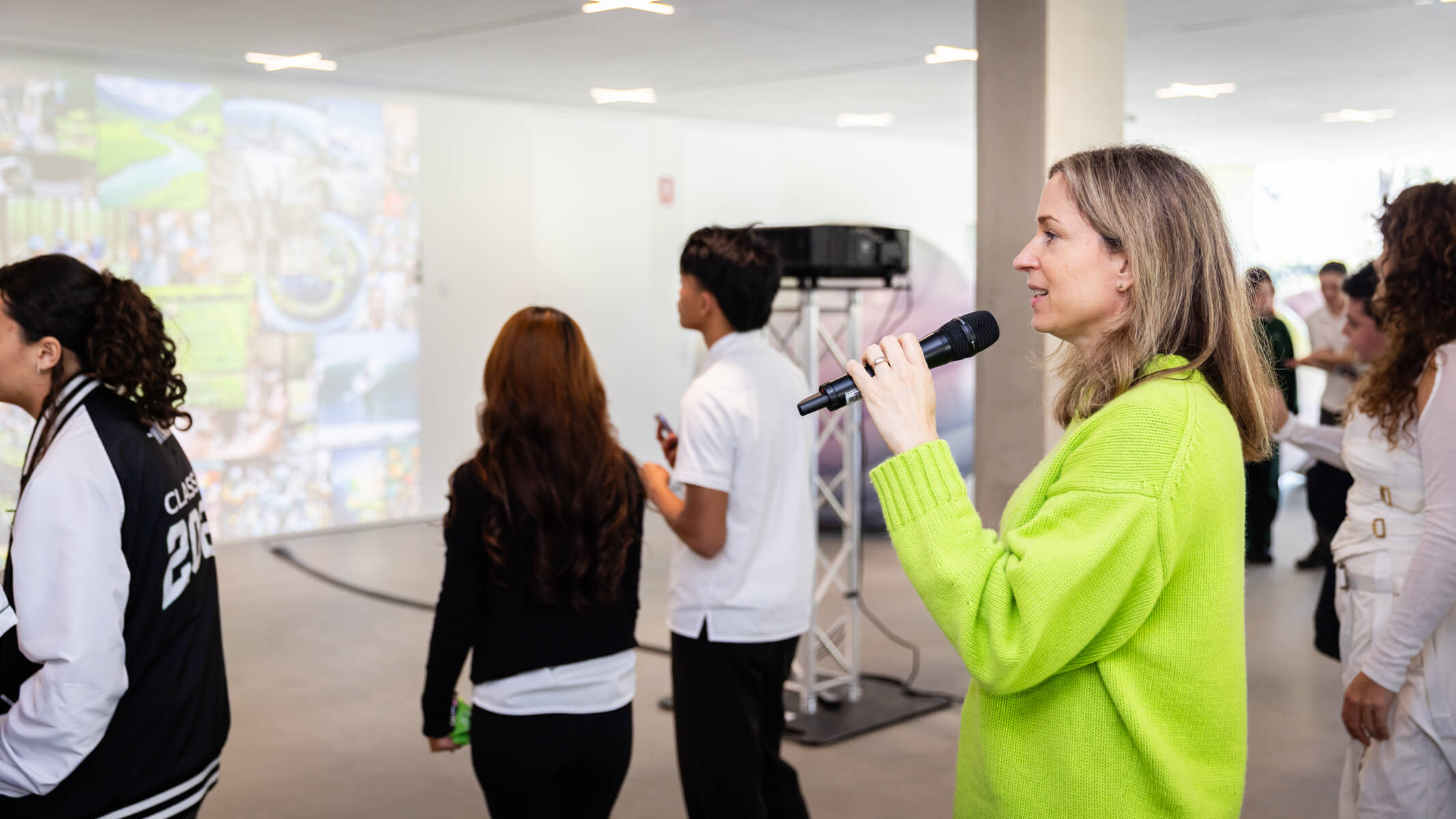 A woman in a green jumper holds a microphone as she speaks to high school students that are dispersed in a white indoor gallery.