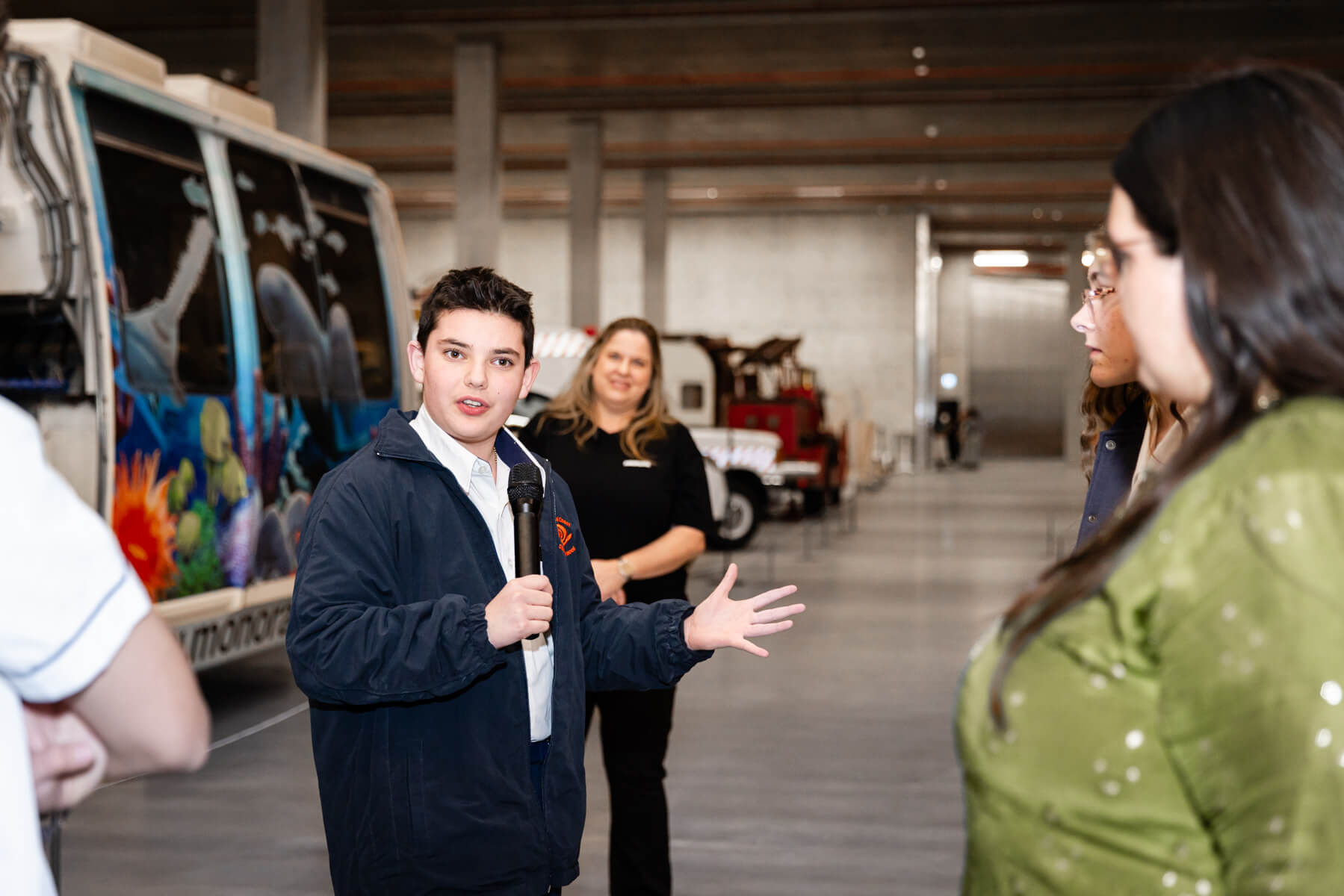 A student stands with a microphone amidst the Very Large and Complex Object storeroom objects at Powerhouse Castle Hill.