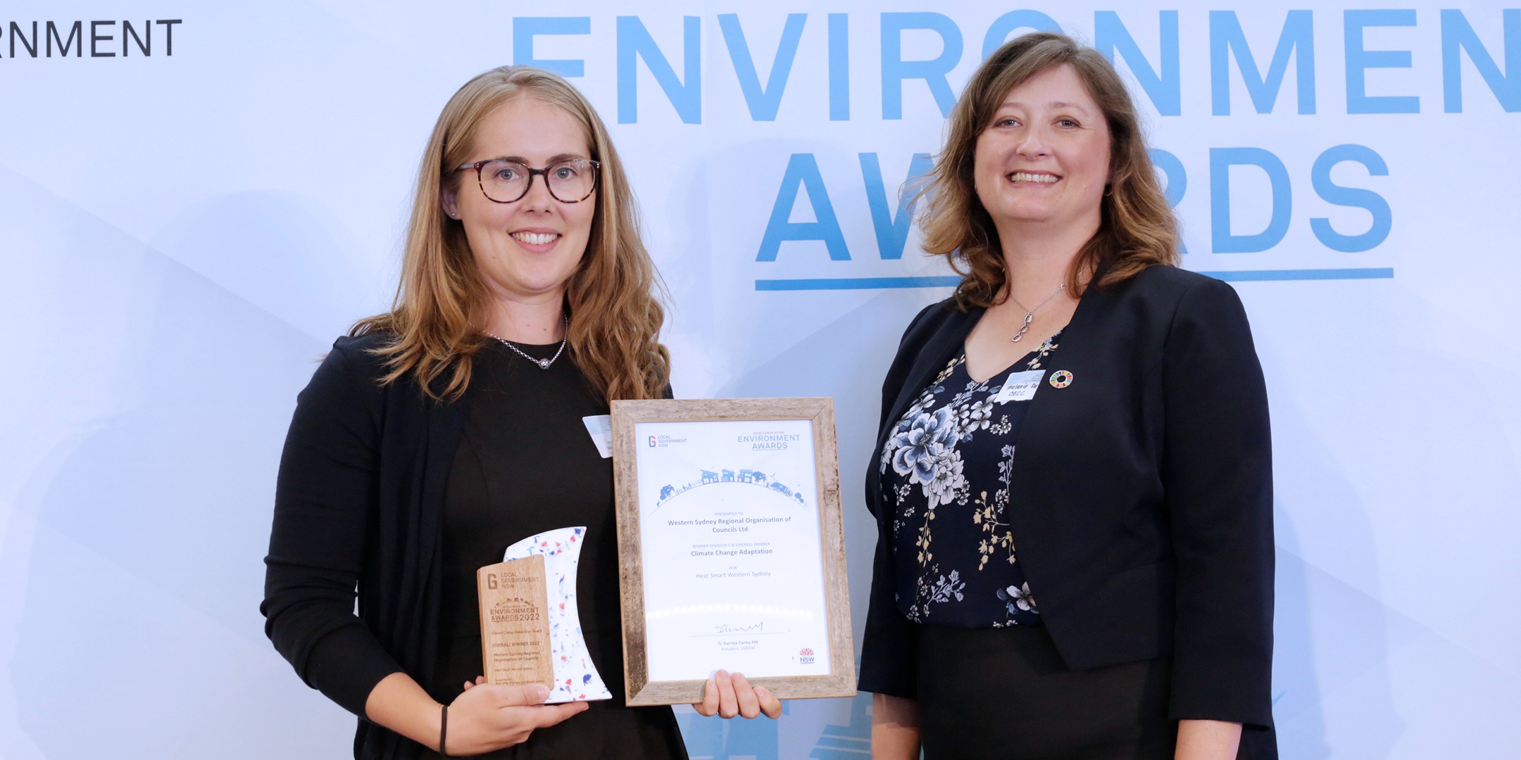 Two people standing side by side, the person on the left is holding an awards and smiling.