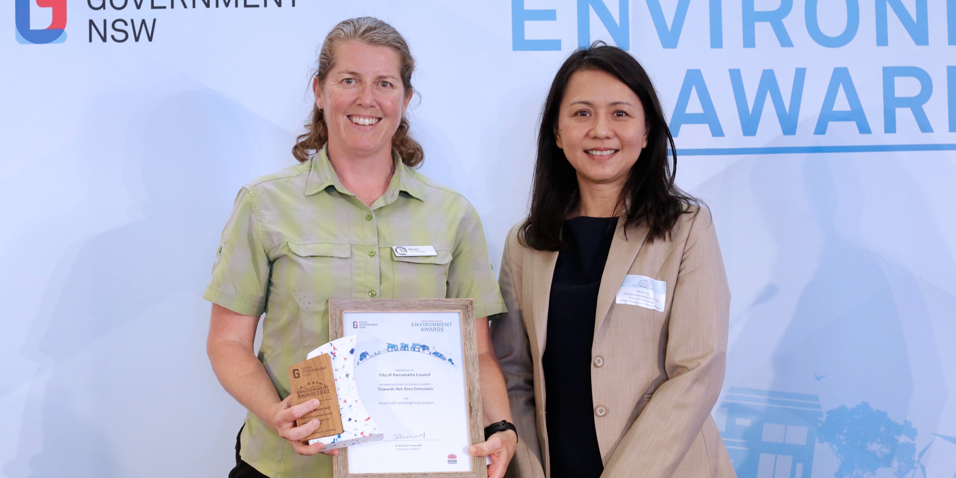 Two people standing side by side, the person on the left is holding an awards and smiling.