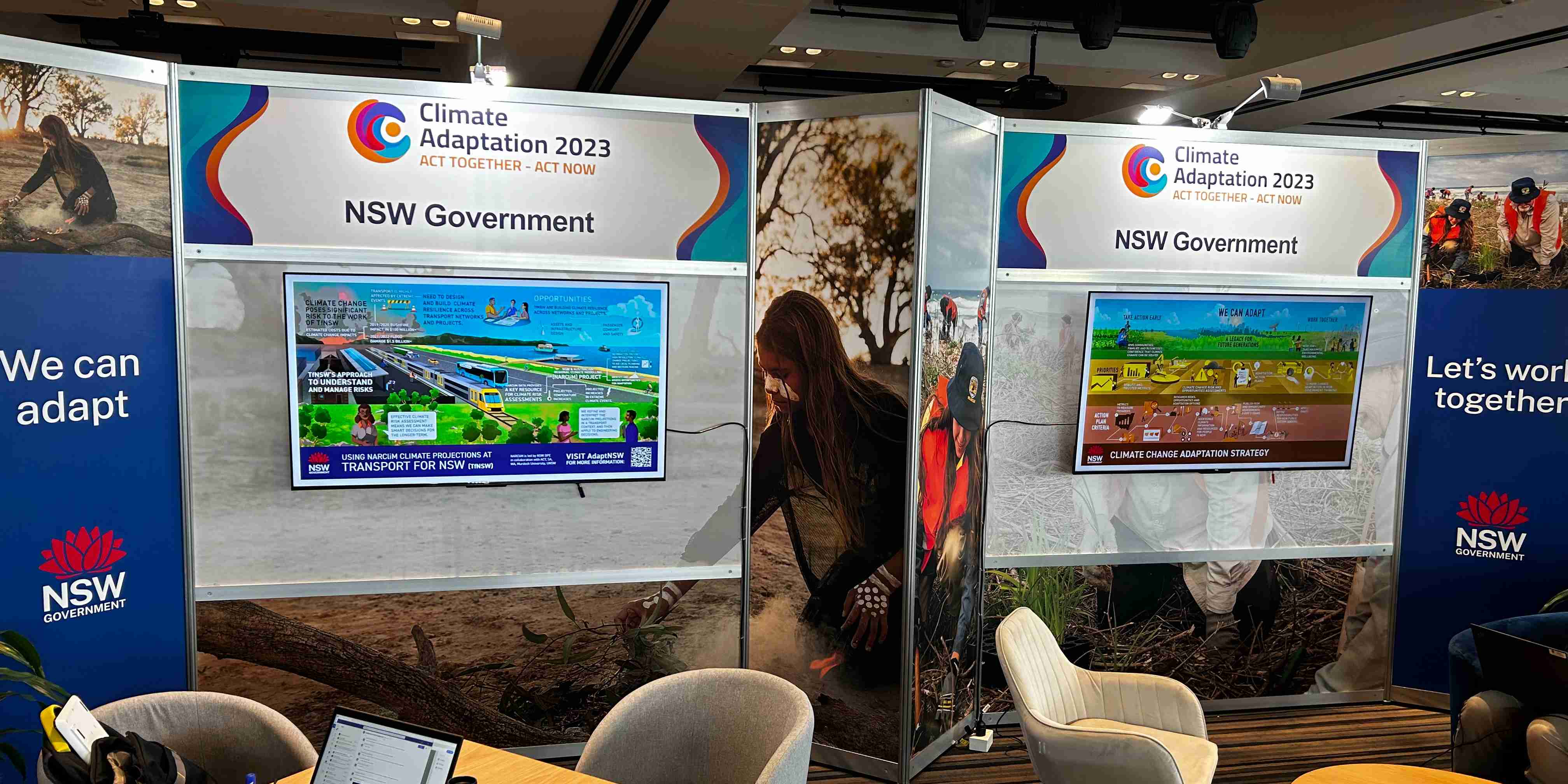 A photograph of the AdaptNSW conference booth. It has two large LCD screens showing AdaptNSW related content and is surrounded by tables and chairs.